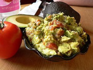 A bowl of guacamole beside a tomato and a cut ...