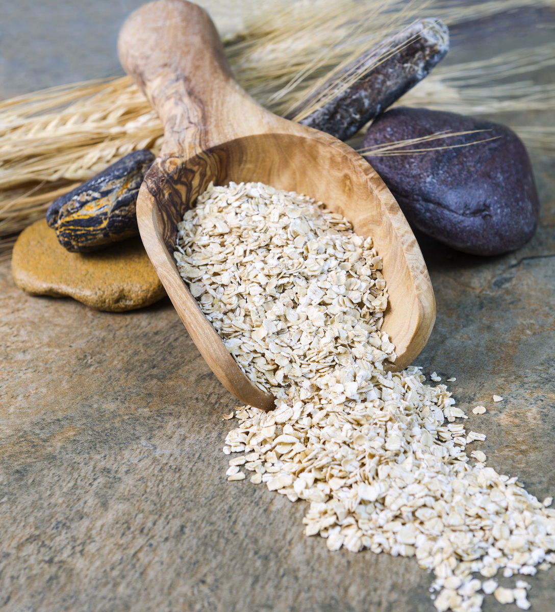 Vertical photo of rolled oats in wooden spoon with rocks and wheat stalks on natural stone